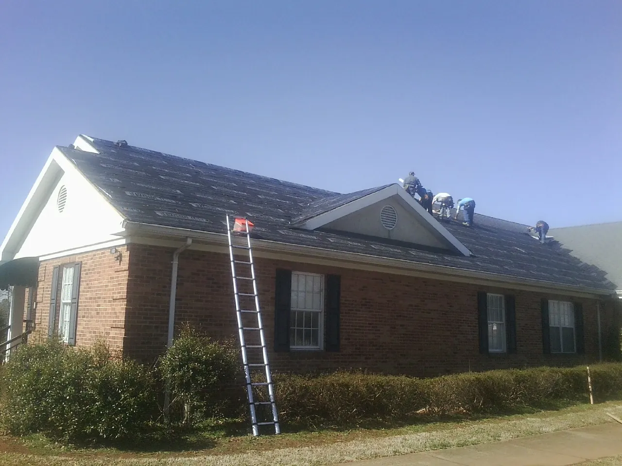 Crew tearing shingles off a brick ranch roof with a ladder set — Emergency Roof Repair work by our crew in Green Camp