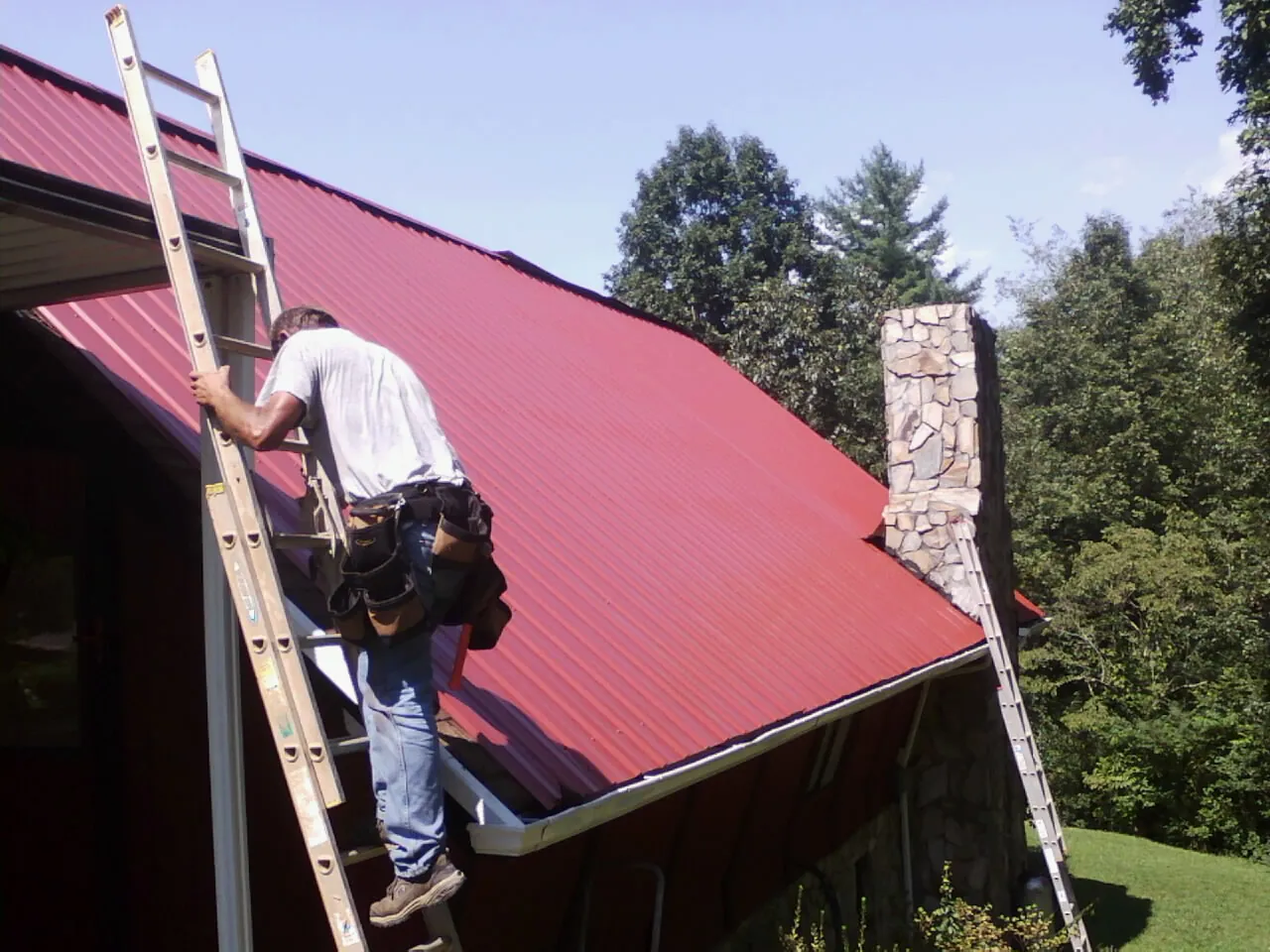 Roofer on a ladder beside a red metal roof and stone chimney — Soffit & Fascia Repair by Roofing Brothers in Green Camp
